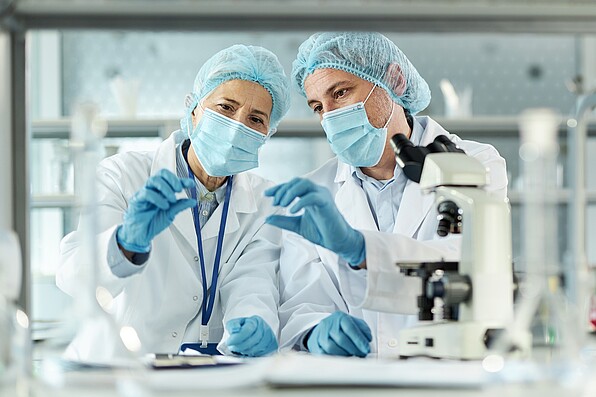 Two scientist with lab coats, masks and hair nets look at a microscope slide. There is a microscope in the front